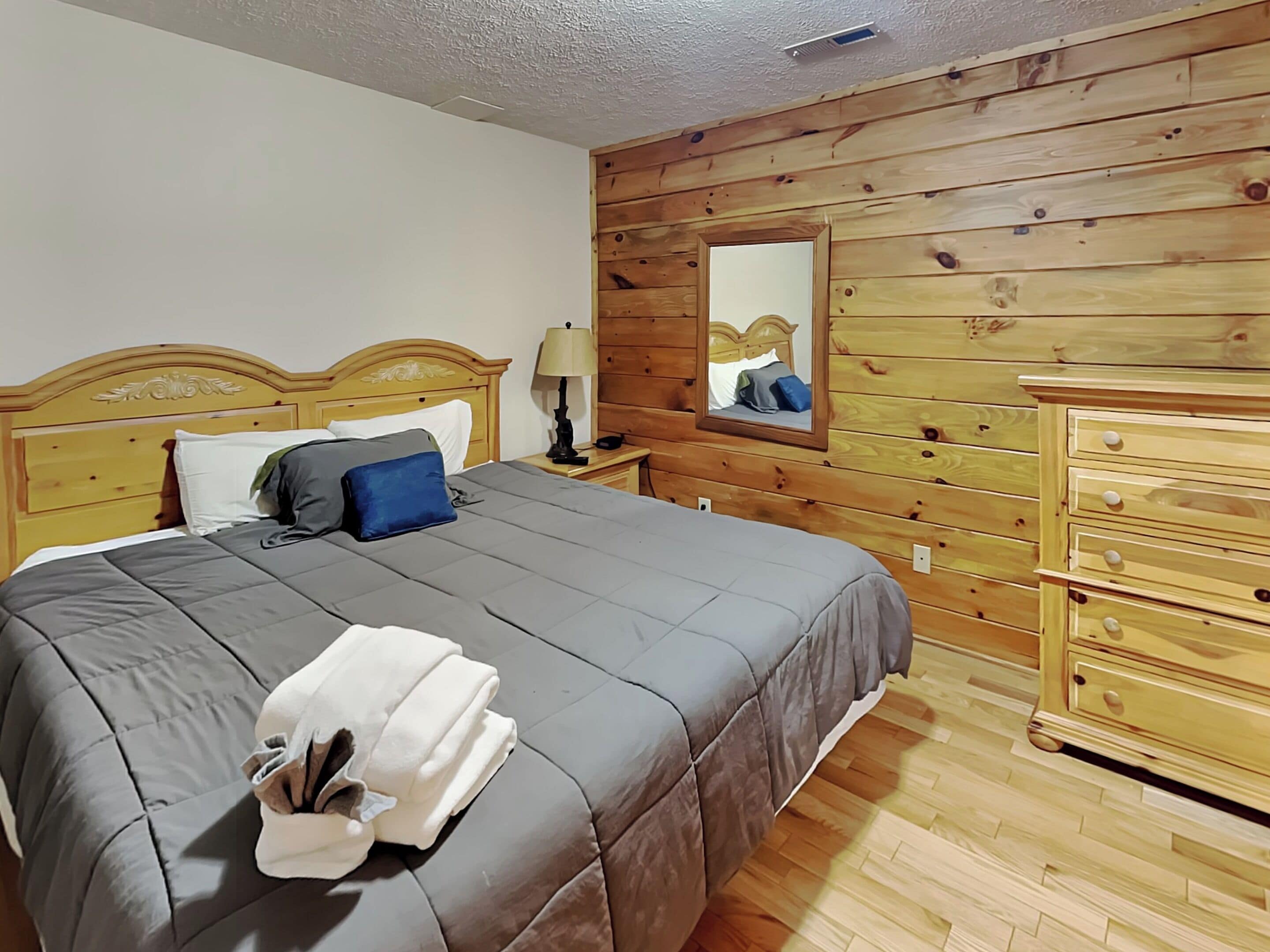 Un dormitorio con cabecera de madera, cama con ropa de cama gris, mesita de noche con lámpara, cómoda y espejo en la pared.