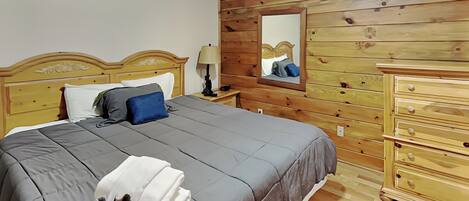 Un dormitorio con cabecera de madera, cama con ropa de cama gris, mesita de noche con lámpara, cómoda y espejo en la pared.