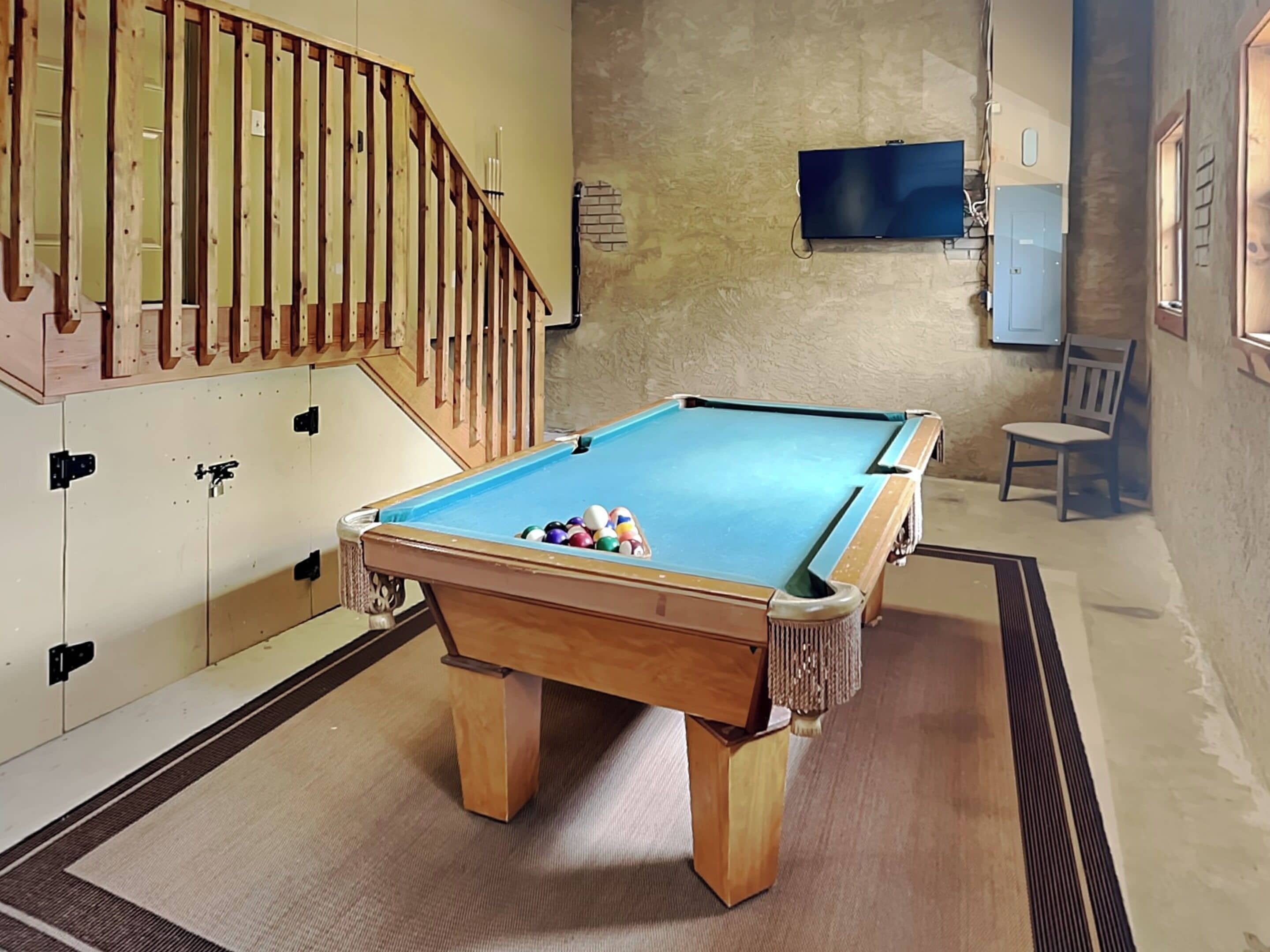 Una mesa de billar con bolas, una escalera de madera, un televisor en la pared y una silla.