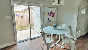 Un comedor con una mesa redonda y cuatro sillas, una araña, un ventanal corredero que da al exterior y un cuadro abstracto colorido en la pared.