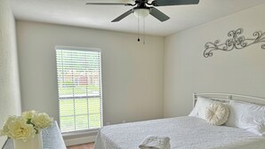 Un dormitorio con una cama, ventilador de techo, ventana con persianas y un adorno en la pared.