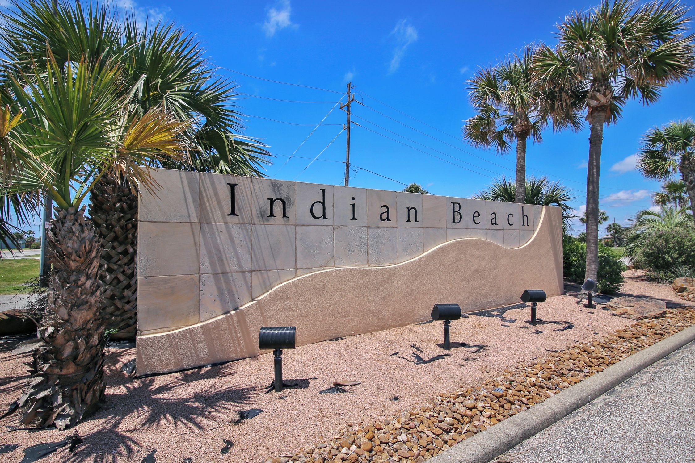 Señal de Indian Beach con un diseño curvo, palmeras y un cielo despejado.