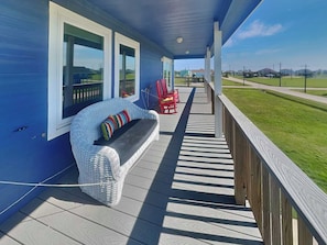 A covered porch with a wicker sofa and red chairs, a wooden deck, and a view of a green landscape. - Splish Splash Oasis by Ryson Vacation Rentals (Galveston)