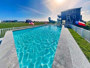 A swimming pool with a white fence, American and Texas state flags, and a house with balconies. - Splish Splash Oasis by Ryson Vacation Rentals (Galveston)