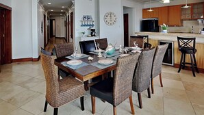 A dining area with a wooden table set for six, surrounded by wicker chairs. A kitchen with wooden cabinets and a countertop is visible in the background. - Sunset On The Seawall #1 by Ryson Vacation Rentals (Galveston)