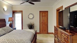 A bedroom with a bed, a dresser, a television, a ceiling fan, a mirror, and a door. - Sunset On The Seawall #1 by Ryson Vacation Rentals (Galveston)