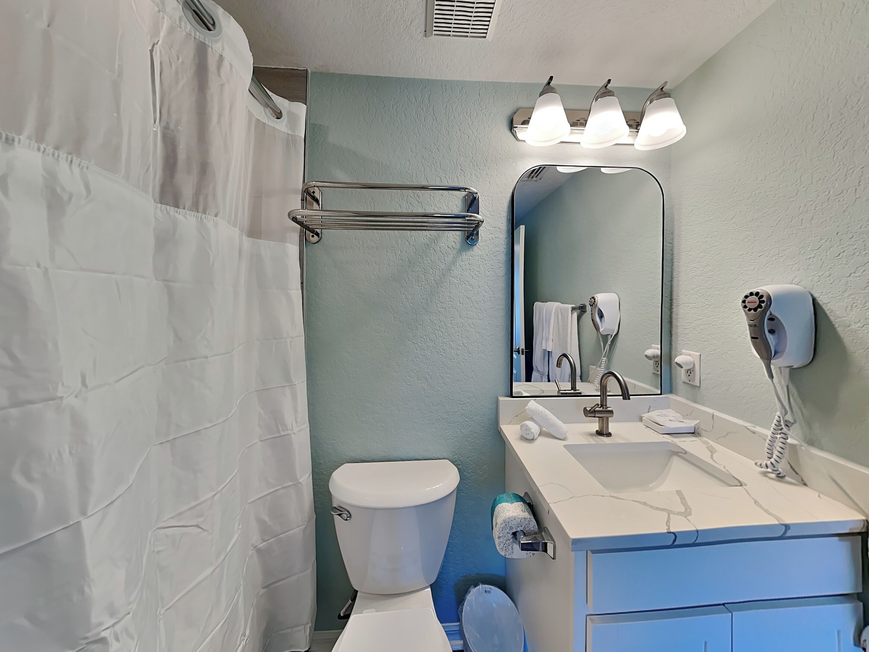 Een badkamer met douchegordijn, toilet, wastafel met spiegel en een aan de muur bevestigde haardroger.