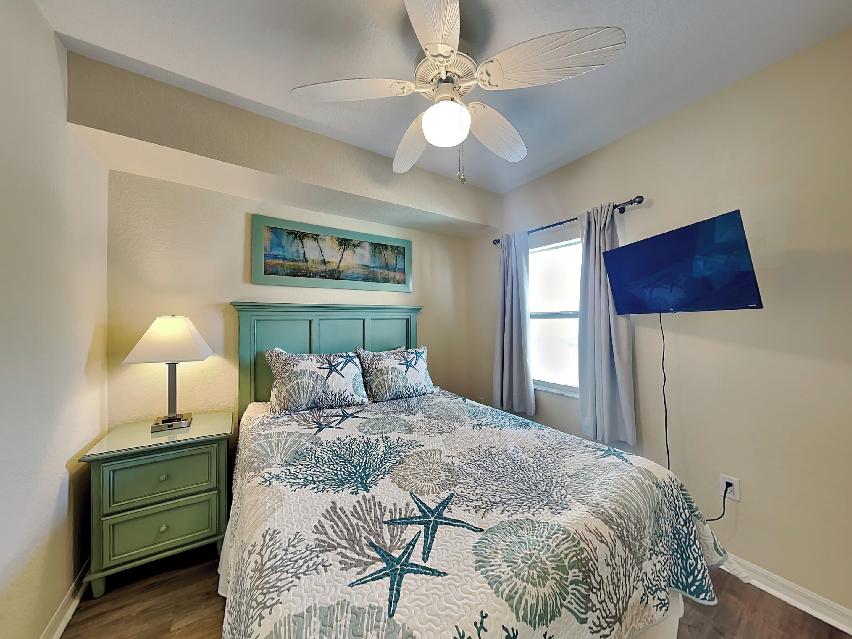 A bedroom with a bed, a nightstand, a ceiling fan, a painting, a window with curtains, and a TV mounted on the wall.
