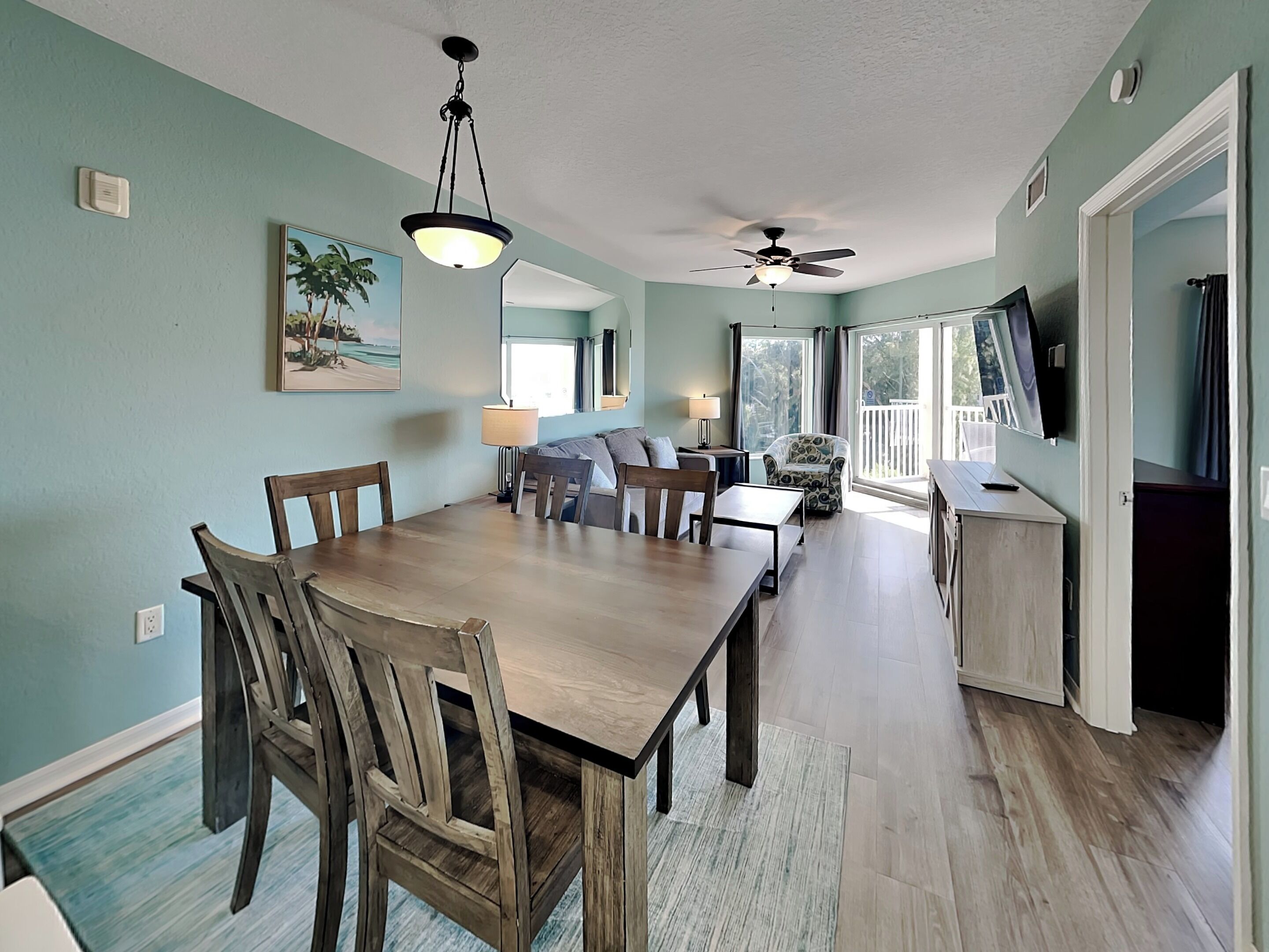 A dining area with a wooden table and chairs, a ceiling fan, and a view of the outdoors through large windows.