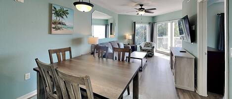 A dining area with a wooden table and chairs, a ceiling fan, and a view of the outdoors through large windows.