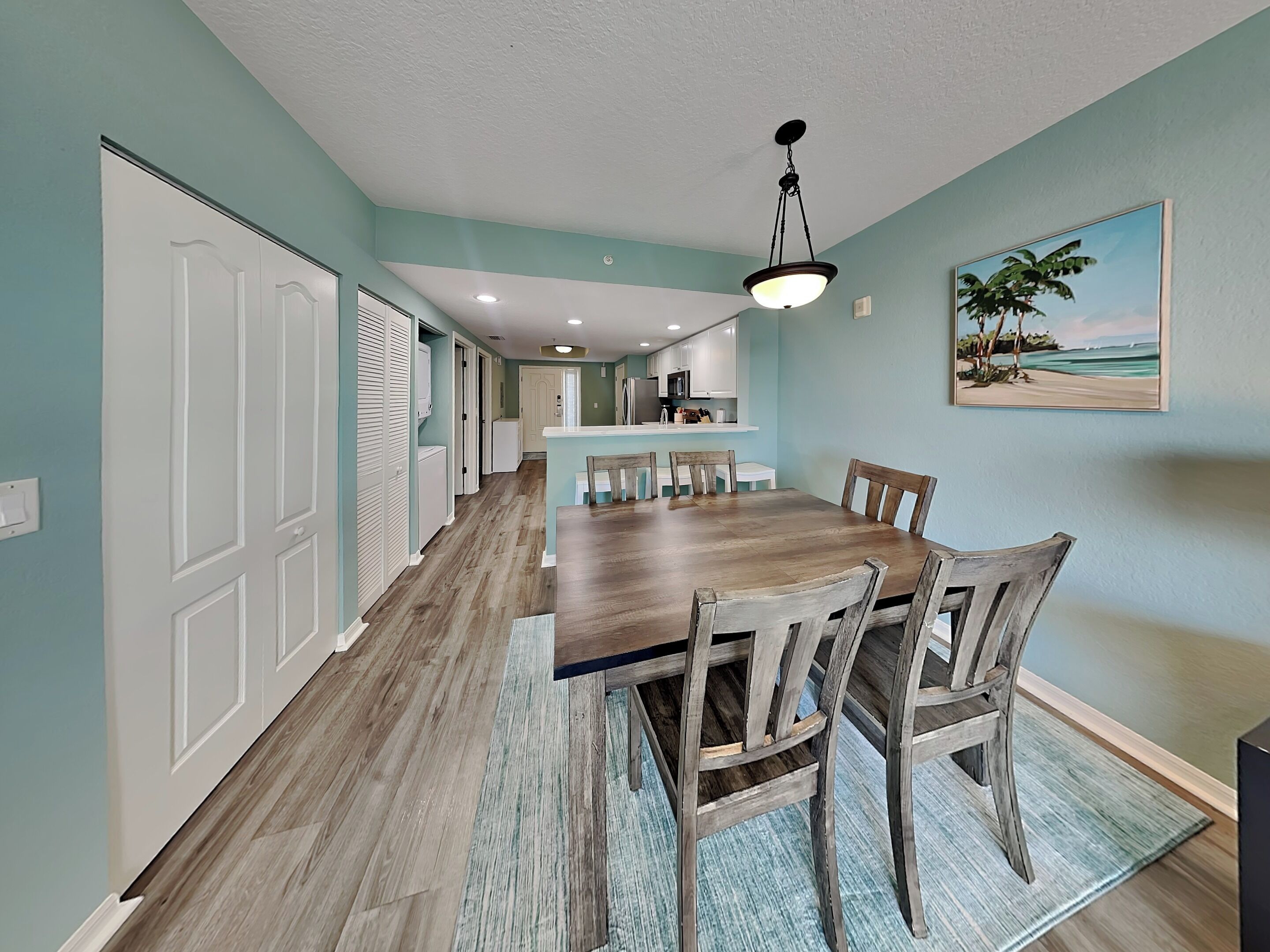 A dining area with a wooden table and chairs, a kitchen in the background, and a painting on the wall.