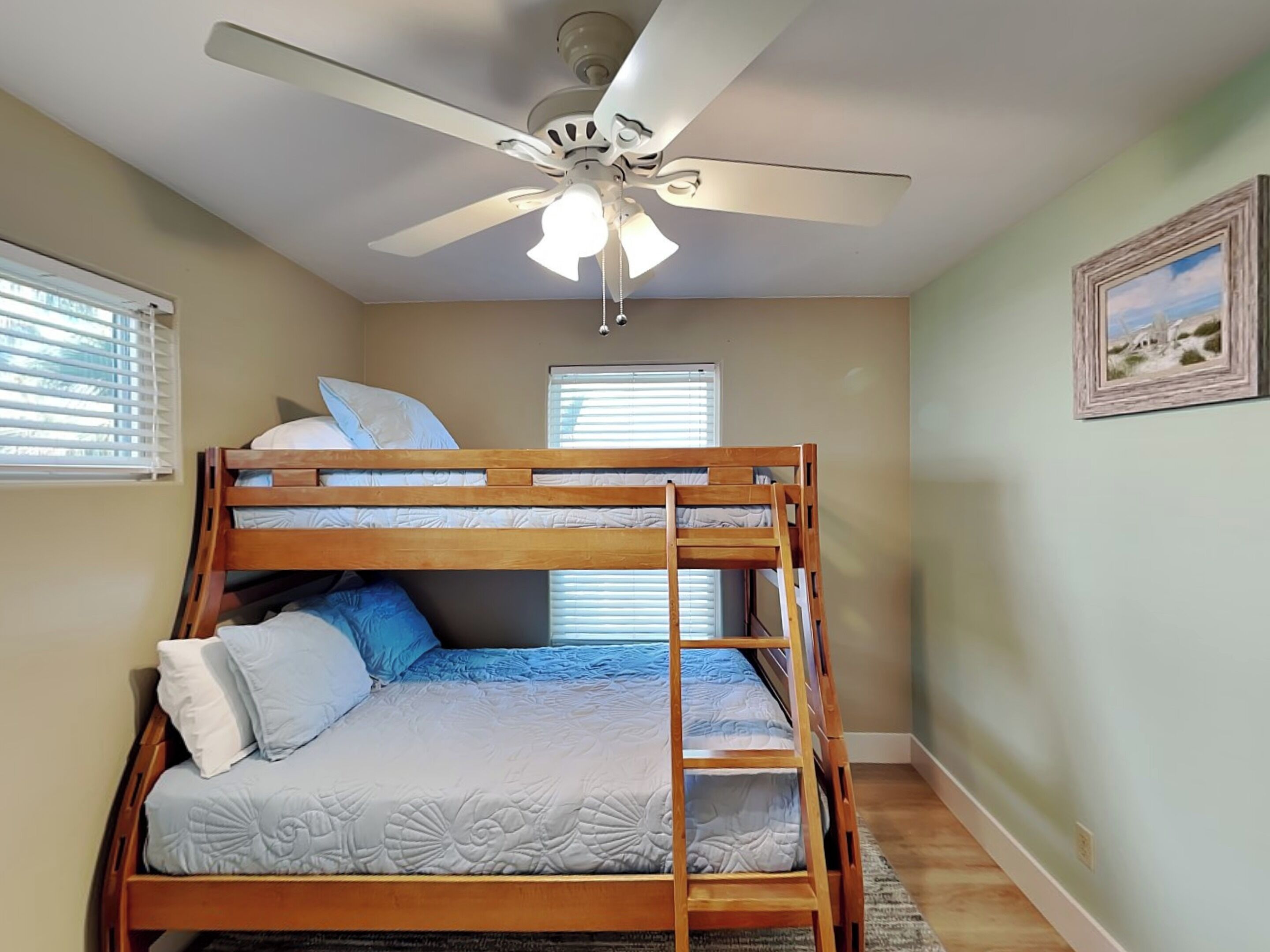 A bunk bed room with a ceiling fan, two windows with blinds, and a framed picture on the wall.
