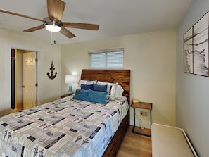 Un dormitorio con cabecera de madera, colcha acolchada, mesita de noche con lámpara, ventilador de techo y ventana con persianas.
