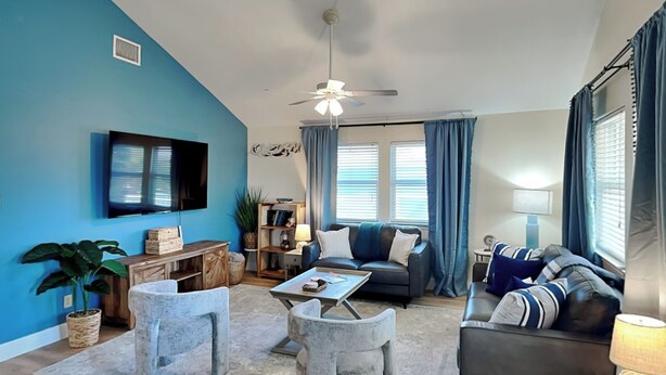 Ein modernes Wohnzimmer mit blauer Akzentwand, einem Flachbildfernseher, einem Deckenventilator und einer gemütlichen Sitzecke mit Sofa und Sesseln.