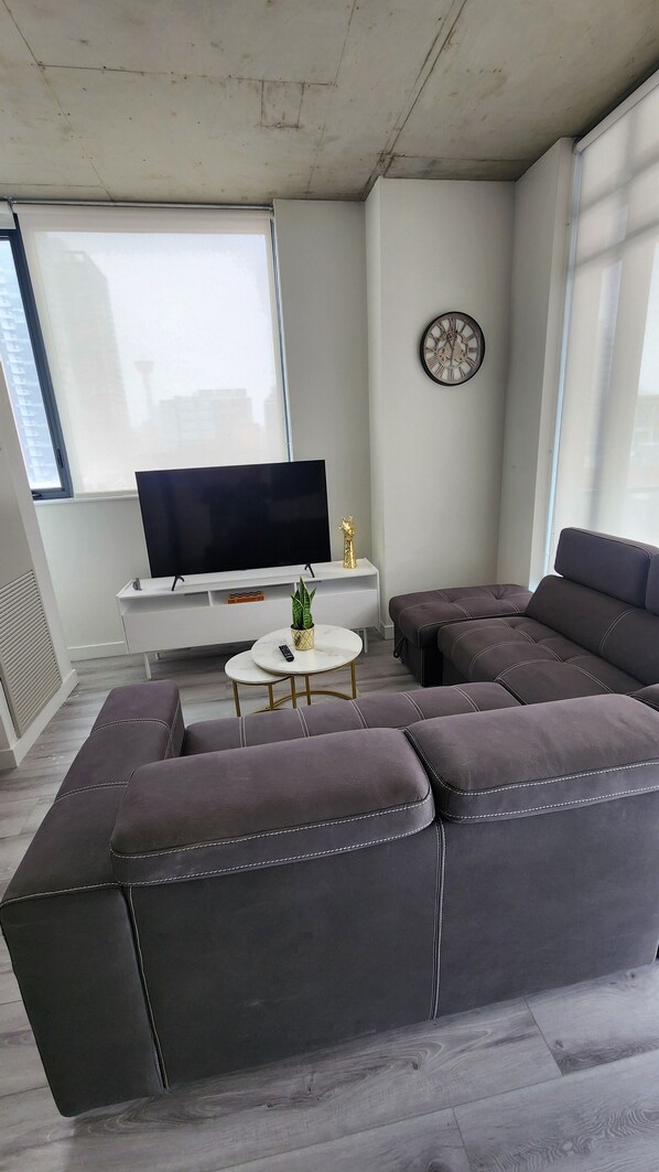 En moderne stue med en hjørnesofa, et fladskærms-tv, et lille rundt sofabord og et dekorativt vægur.
