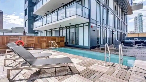 A rooftop pool area with lounge chairs, a wooden deck, and a glass building in the background.