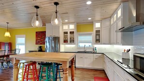A modern kitchen with white cabinets, a stainless steel refrigerator, and a wooden dining table with colorful stools. - The Reel Mccoy by Miss Kitty’s (Rockport)