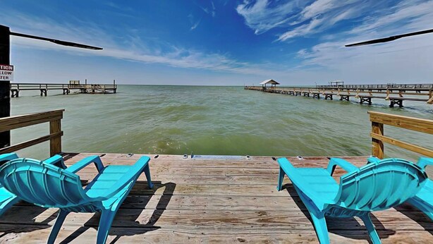 A wooden pier with two blue chairs facing the ocean, a sign warning of shallow water, and a distant pier extending into the sea. - The Reel Mccoy by Miss Kitty’s (Rockport)