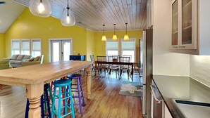 A spacious kitchen with wooden flooring, a central island with stools, pendant lighting, and a large window. - The Reel Mccoy by Miss Kitty’s (Rockport)