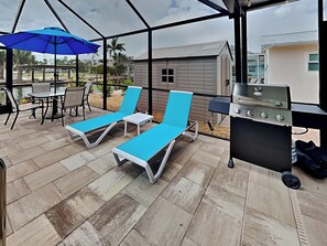 A covered patio with blue lounge chairs, a grill, and a table with chairs.