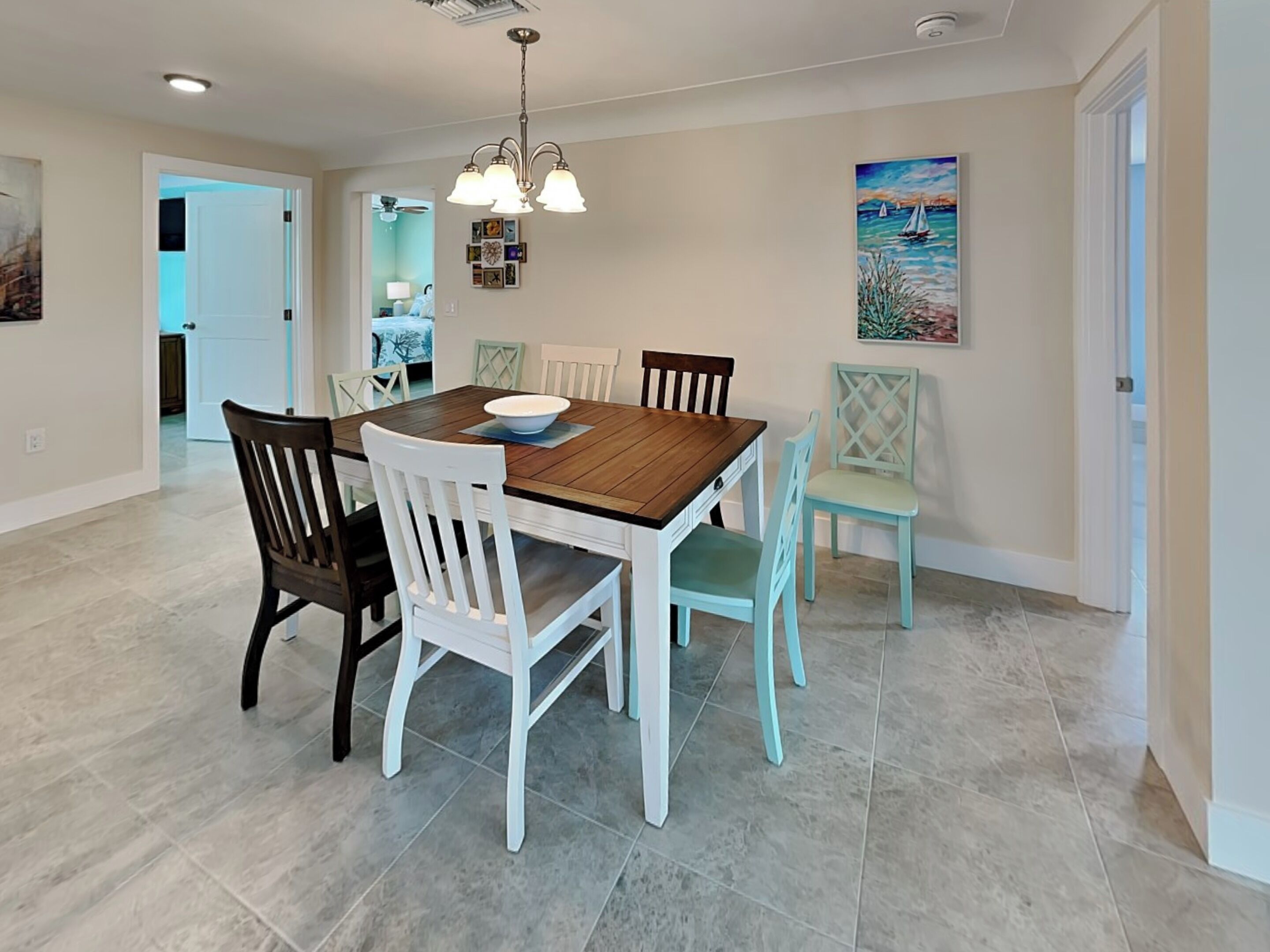 A dining room with a wooden table, white and light blue chairs, a chandelier, and a painting on the wall.