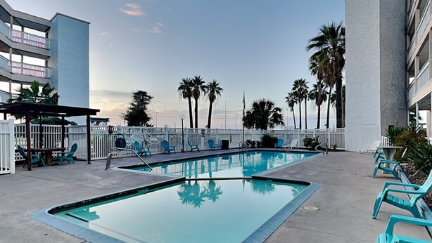 A swimming pool with lounge chairs and a fenced area, surrounded by palm trees and a building. - Villa Del Sol #2138 by Miss Kitty’s (Corpus Christi)