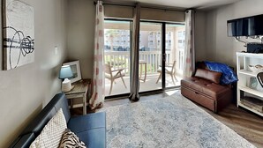 A room with a ceiling fan, a sofa, a TV, and a balcony with a view of other buildings. - Villa Del Sol #2138 by Miss Kitty’s (Corpus Christi)