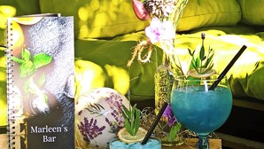 Un libro con el título 'Marleen's Bar' sobre una mesa, con dos bebidas azules y un arreglo floral.