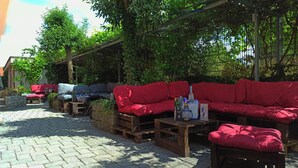 Un área de estar al aire libre con varios sofás rojos y mesas de madera, rodeada de follaje y una estructura de pérgola.