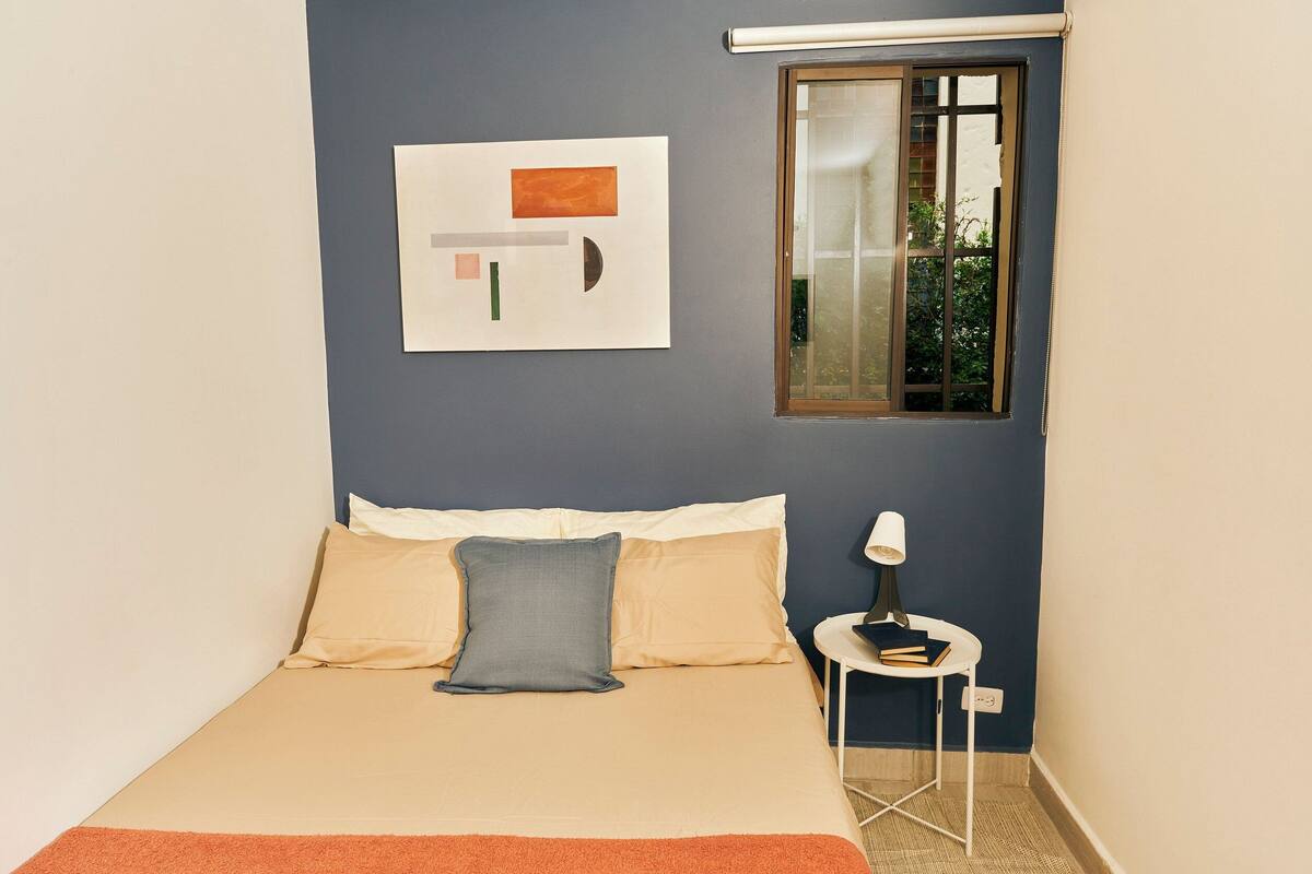 A bedroom with a bed, pillows, a bedside table with a lamp, a window, and a wall art piece.