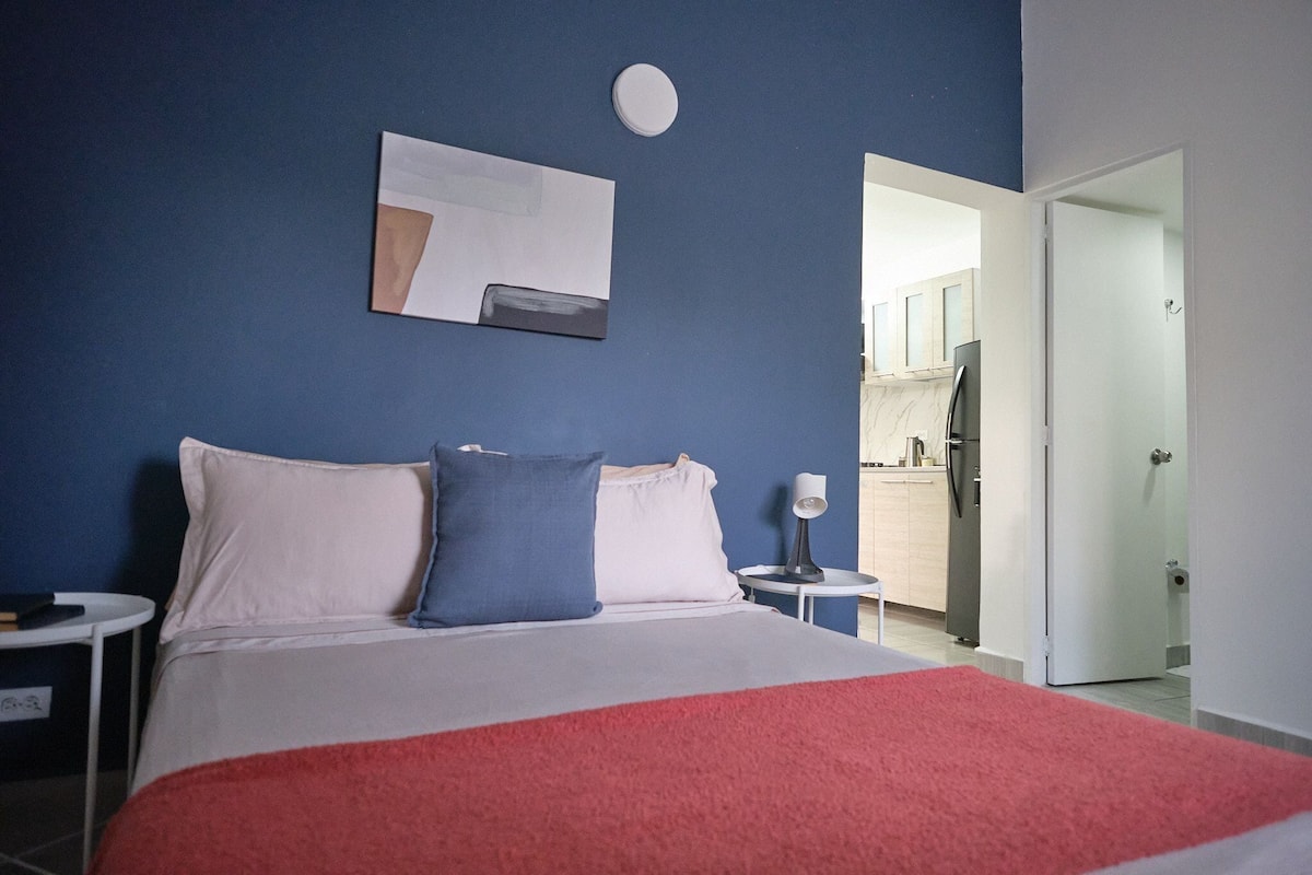 A bedroom with a bed, pillows, a bedside table, a wall art piece, and a view into the bathroom.