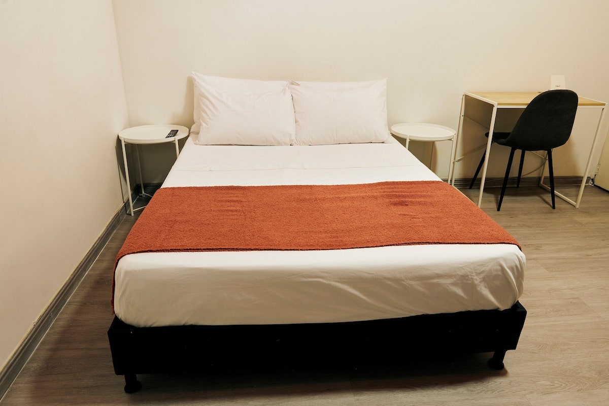 A neatly made bed with white linens and a brown throw, a small round table with a phone, and a desk with a chair in a room with light-colored walls and flooring.