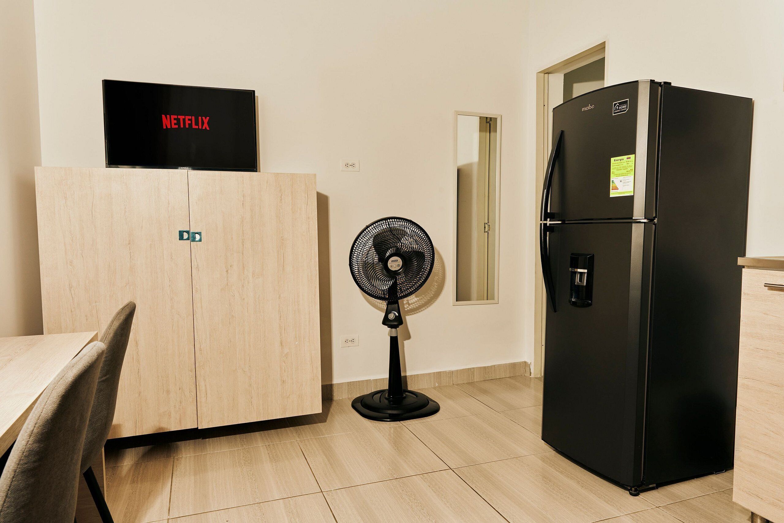 Una kitchenette con un refrigerador negro, un armario de madera, un ventilador de pie y un televisor mostrando el logotipo de Netflix.