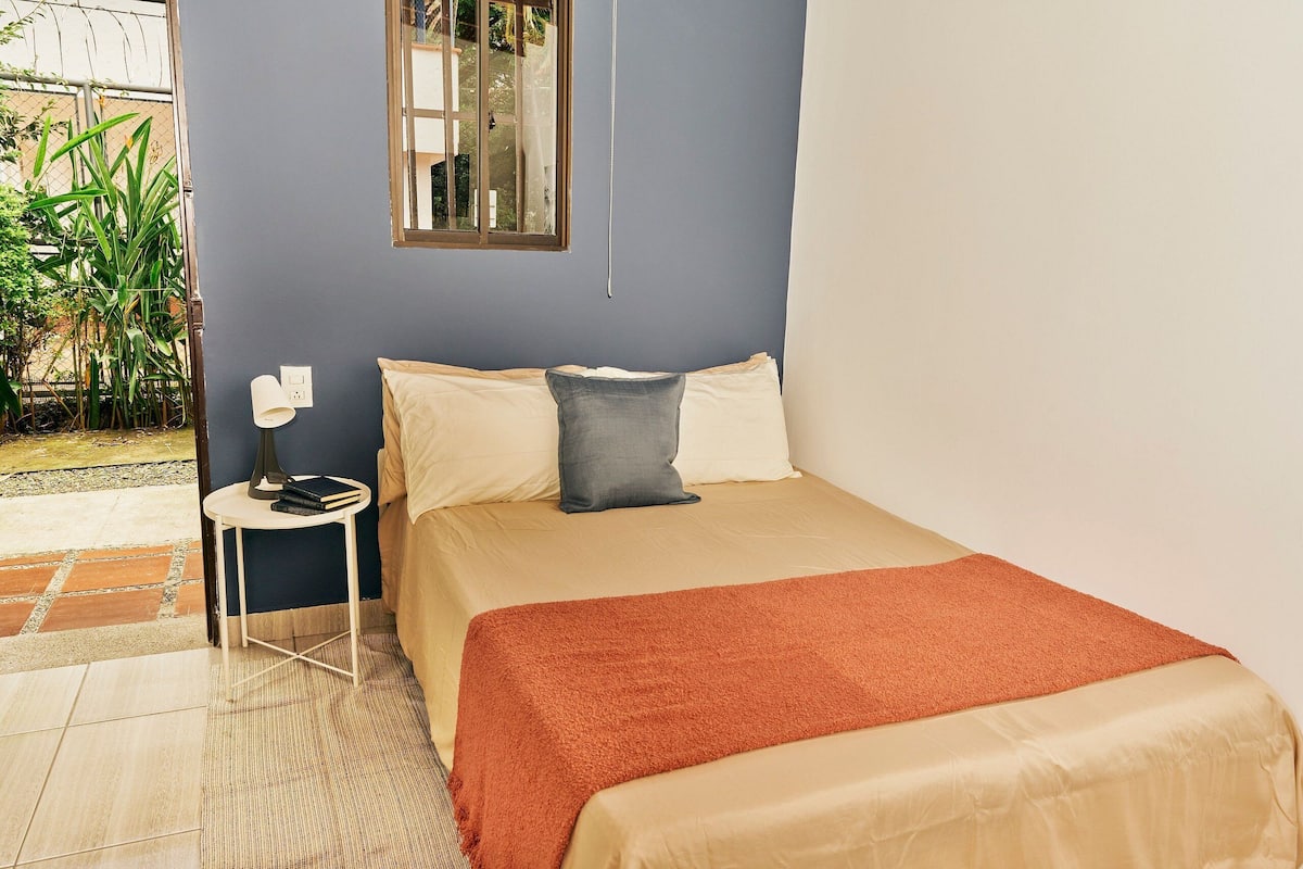 A neatly made bed with a beige and orange blanket, a small bedside table with a lamp, and a window with a view of greenery outside.