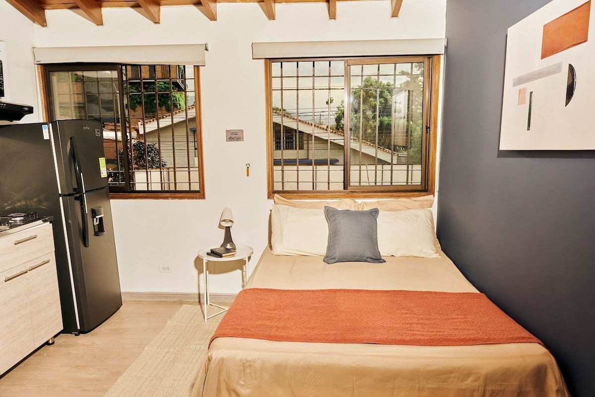 A bedroom with a bed, a nightstand, a lamp, a window with a view of a staircase, and a refrigerator.