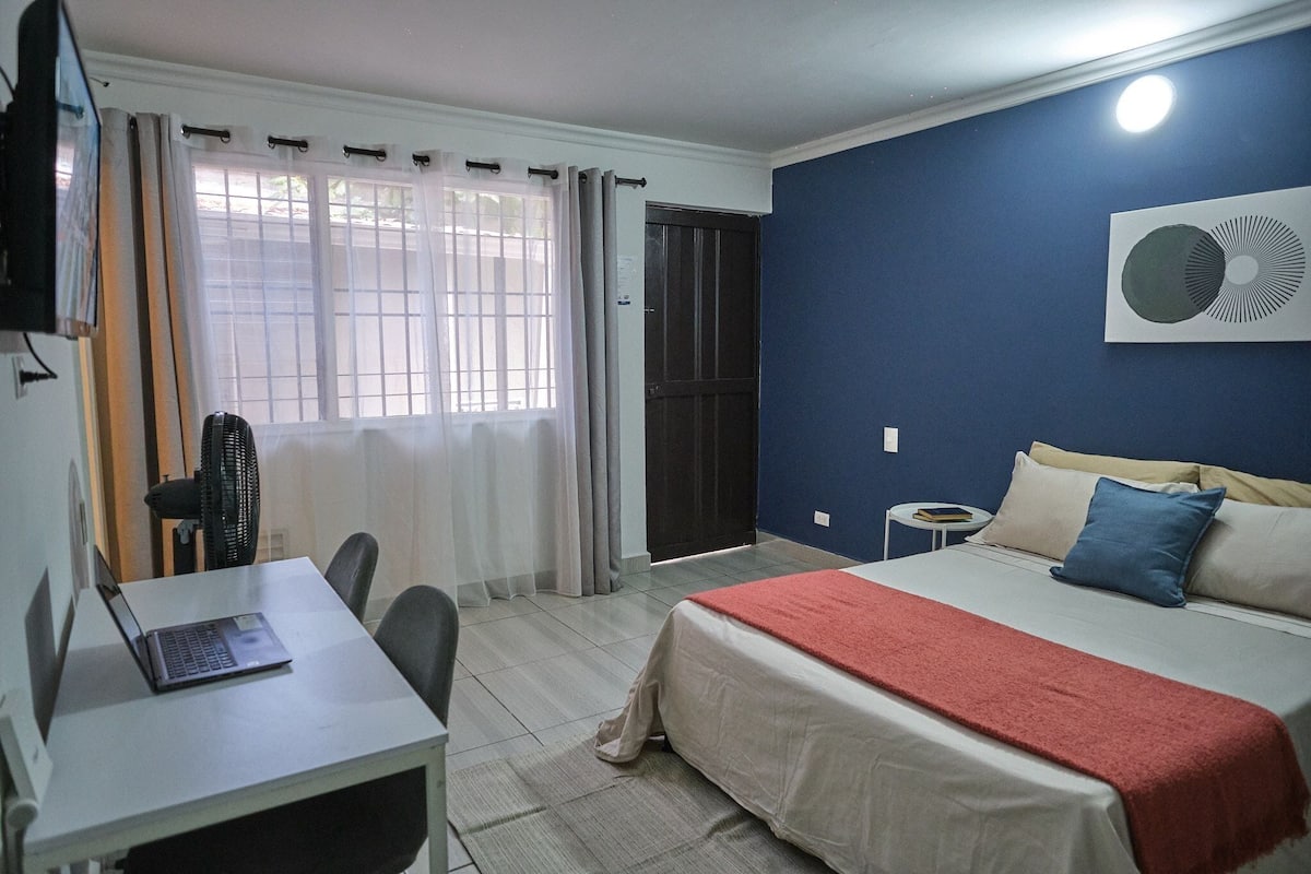 A hotel room with a bed, a desk with a laptop, a chair, a fan, and a window with curtains.