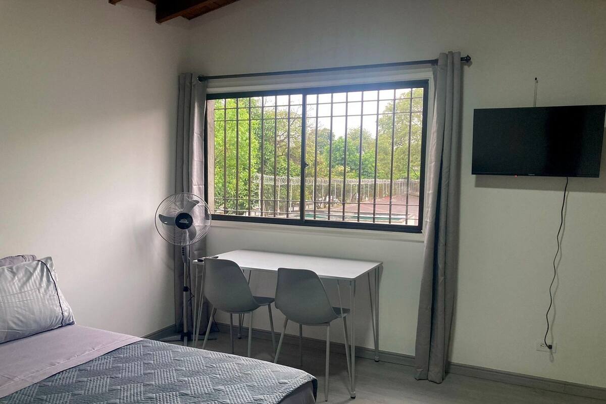 A room with a bed, a desk with two chairs, a standing fan, a TV, and a window with bars.