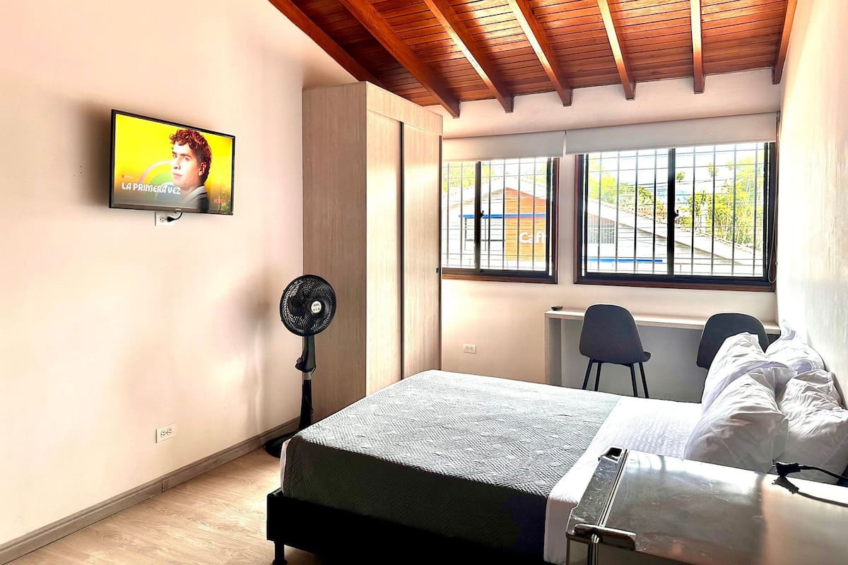 A bedroom with a bed, a desk, two chairs, a TV, and a window with a view of a building.