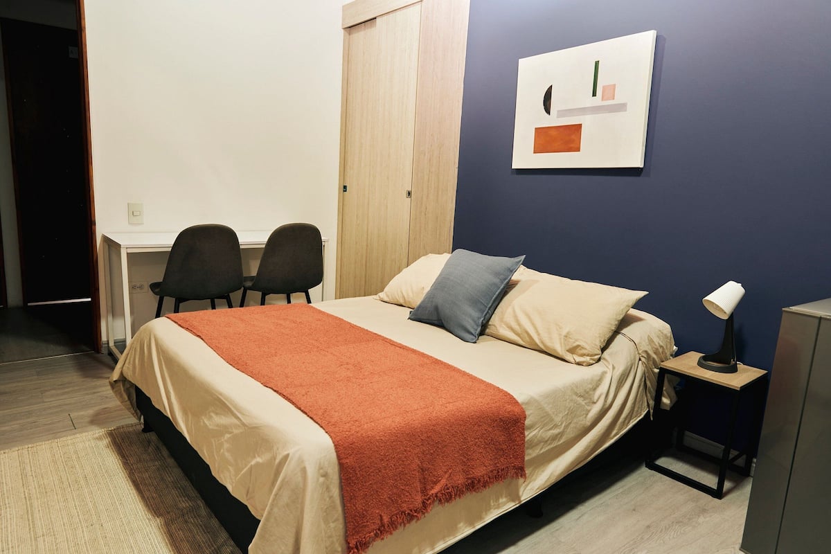 A bedroom with a bed, two chairs, a desk, and a nightstand with a lamp.
