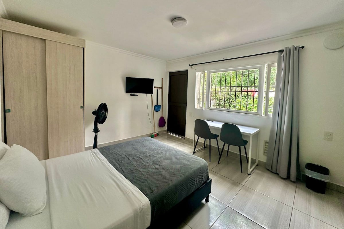 A bedroom with a bed, a desk, two chairs, a TV, and a window with curtains.