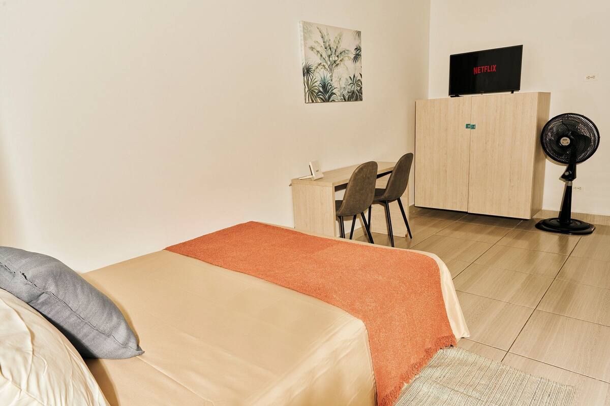 A bedroom with a bed, a desk, a chair, a TV, and a standing fan.