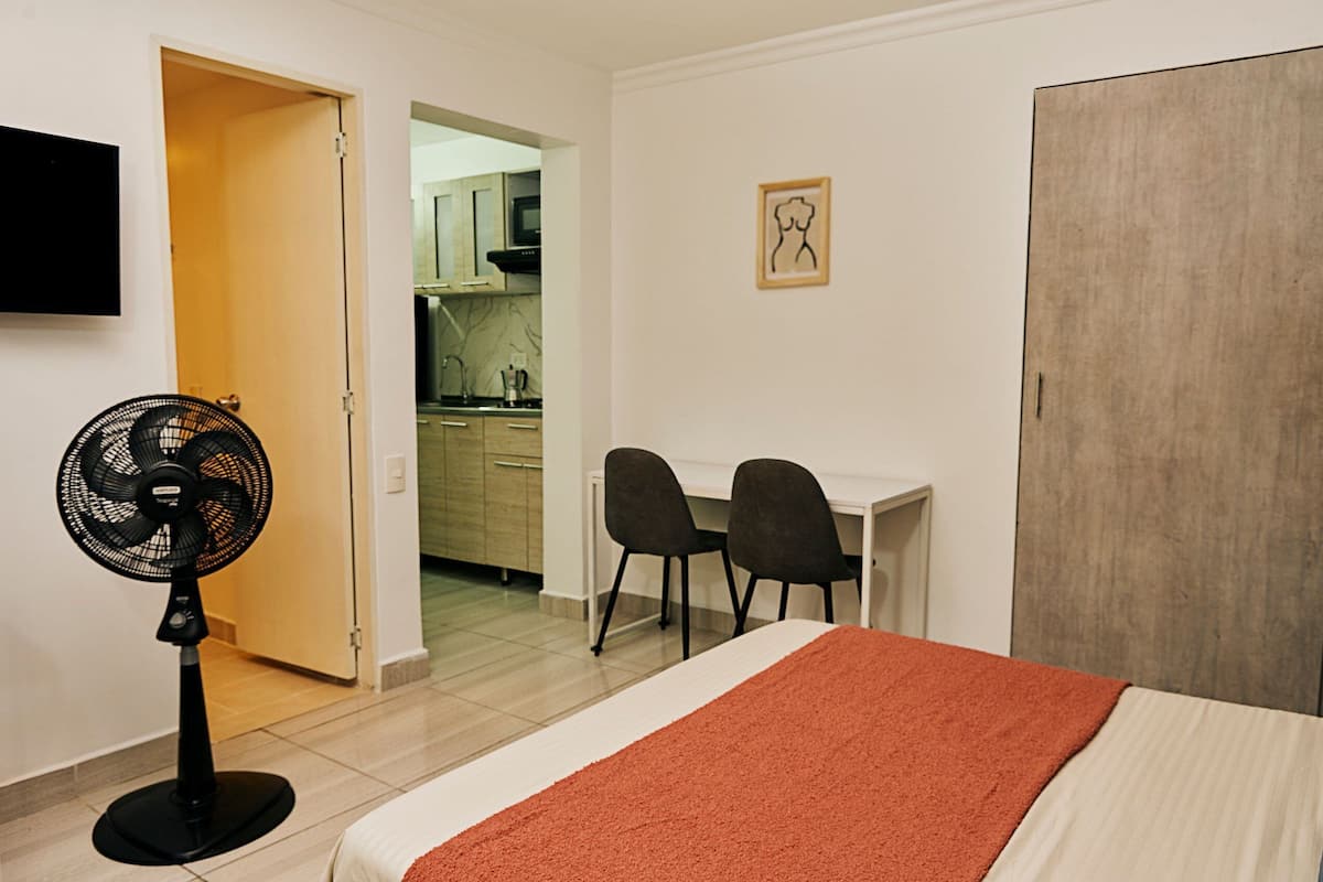 A hotel room with a bed, a desk, two chairs, a standing fan, and a view into the kitchen.