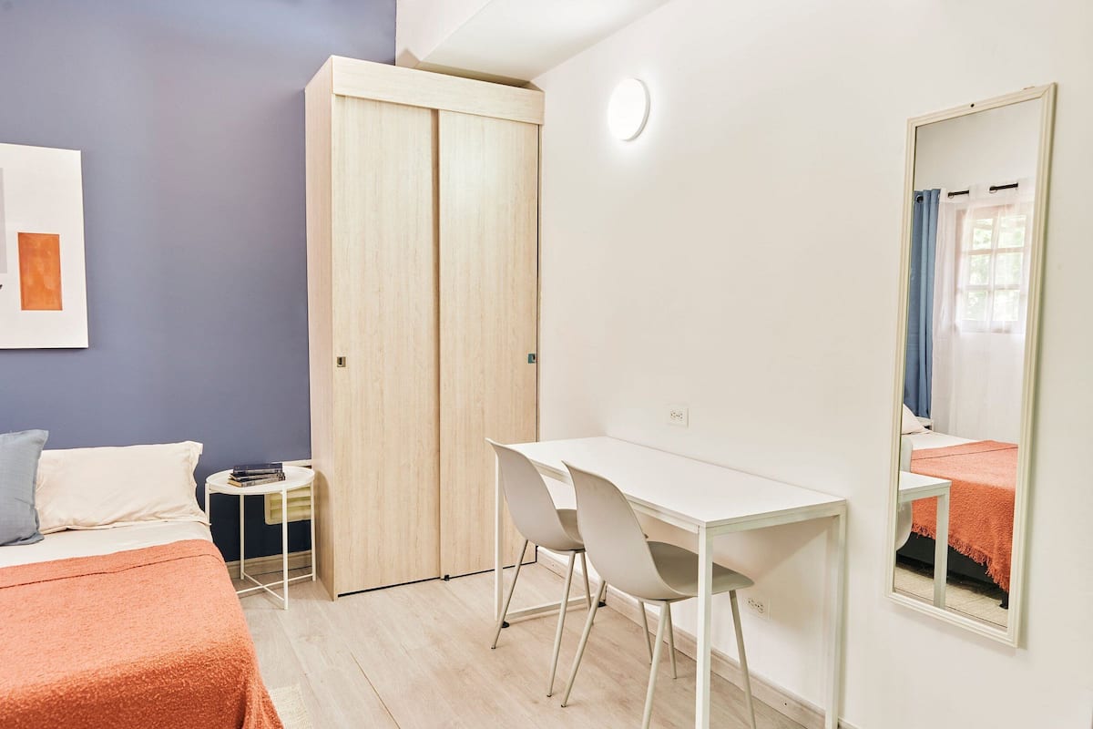 A bedroom with a bed, a desk, two chairs, a wardrobe, a small table, and a mirror.
