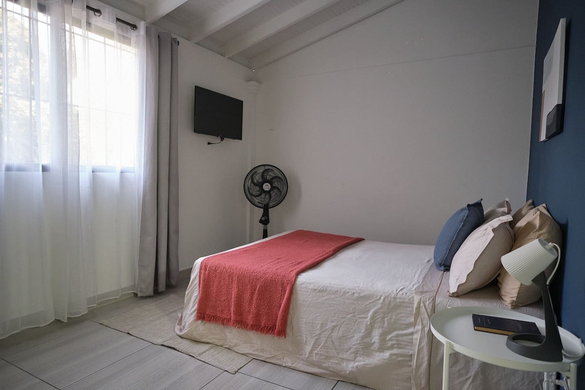 A neatly arranged bedroom with a bed, a bedside table with a lamp, a fan, and a TV mounted on the wall.