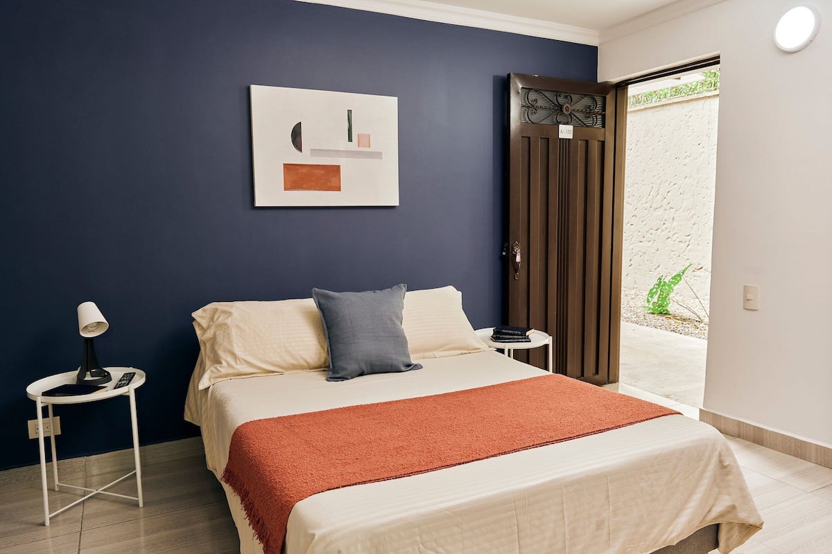 A bedroom with a bed, a bedside table with a lamp, a nightstand, a door, and a window.