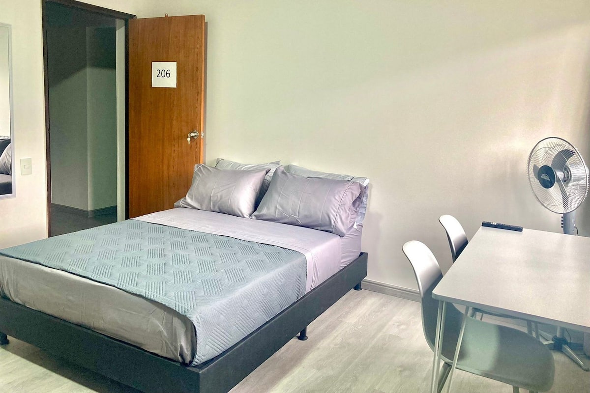 A room with a bed, a table, a chair, a fan, and a door with a number sign.