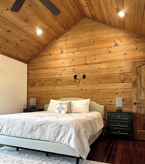 A bedroom with a sloped wooden ceiling, a bed with white bedding, two bedside tables with lamps, and a dark wooden dresser. - Cozy Cabin 2 Miles from Downtown Blue Ridge! (Blue Ridge)