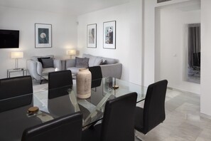 A modern living room with a glass-top dining table, grey sofa, and framed artwork on the walls.
