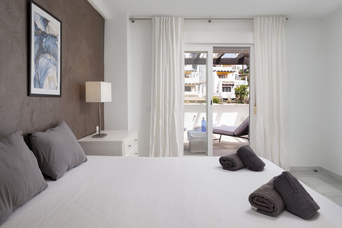 A hotel room with a bed, a nightstand, a lamp, and a view of the outside.