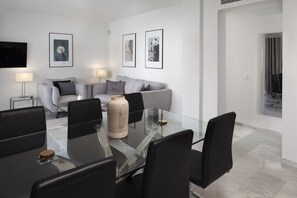 Uma sala de estar moderna com uma mesa de jantar de vidro, sofá cinza e cadeiras pretas. Há quadros nas paredes e uma TV de tela plana montada acima do sofá.